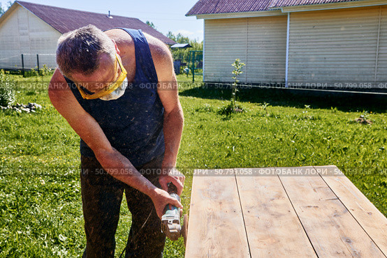 Holding angle grinder with both hands, craftsman wearing respirator and safety glasses sanding tabletop outdoors in rustic countryside setting. Стоковое фото, фотограф Владимир Григорьев / Фотобанк Лори
