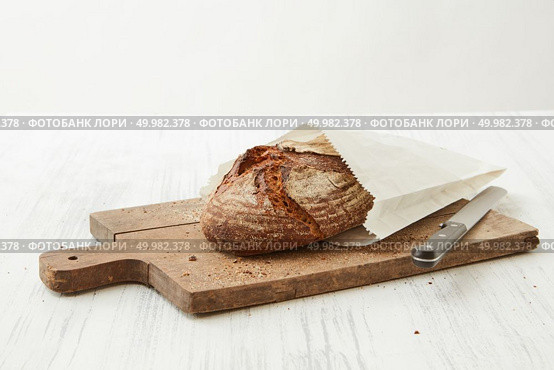 Homemade bread wrapped in paper on a wooden board with a knife on a light background. Fresh oval organic bread. Стоковое фото, агентство Ingram Publishing / Фотобанк Лори