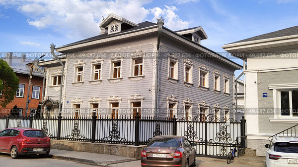 House with carved architraves, cornices and other facade details on Vorovsky Street. Two story building behind blind wrought iron fence. Cars parked along the road. Russia, Vologda, 05.23.2025. Редакционное фото, фотограф Ирина / Фотобанк Лори