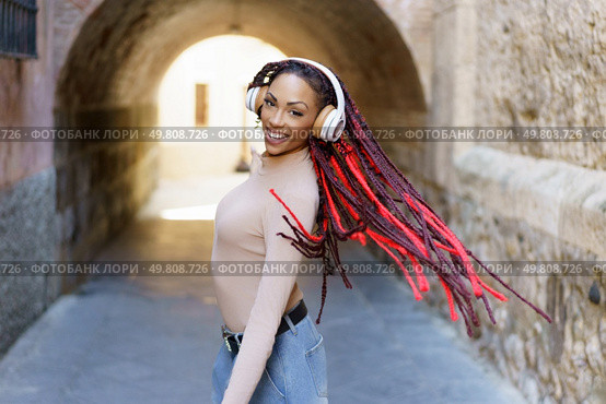 In a lively urban setting, a stylish woman with vibrant red dreadlocks dances gracefully in her headphones, radiating joy, freedom, and confidence as she moves to the musics rhythm. A woman with striking red dreadlocks is enjoying music while standing in an urban archway. Стоковое фото, агентство Ingram Publishing / Фотобанк Лори