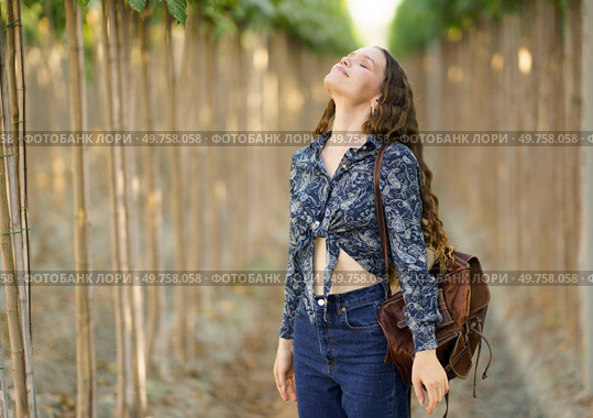 In a sunny vineyard, a young woman stands gracefully among greenery, joyfully embracing nature. Dressed stylishly with a chic brown bag, she radiates tranquility and happiness, a perfect photo moment. A Young Woman Happily Enjoying the Beauty of Nature within a Stunning Vineyard Setting. Стоковое фото, агентство Ingram Publishing / Фотобанк Лори