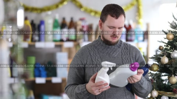 In Christmas decorated shop, man examines product and doubts, chooses laundry fabric softener and and tile cleaner spray, study items properties in household department. Стоковое видео, видеограф Яков Филимонов / Фотобанк Лори