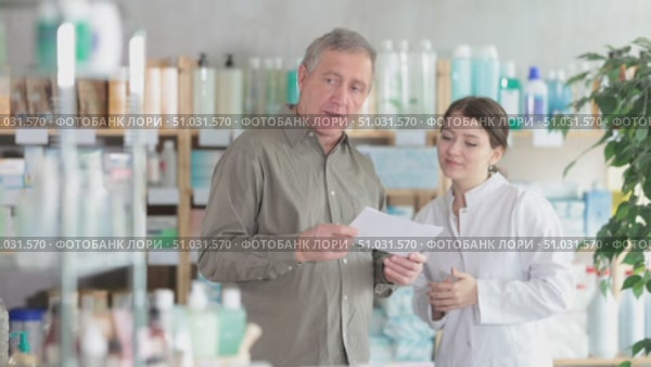 In sales area of drugstore, mature man with prescription intently looks at display cases, trying to find vitamin for adults in open display. Girl pharmacist offer help to customer. Стоковое видео, видеограф Яков Филимонов / Фотобанк Лори