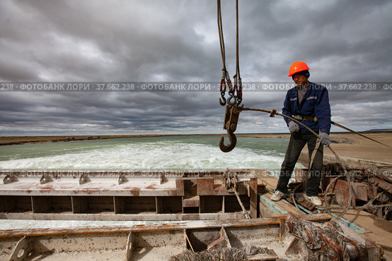 Kok-aral, Kazakhstan - May 03, 2012: Small Aral Sea Kok-aral dam. Young Asian worker lifts water shutter weighting agent. Steel rope, crane hooks. Stormy sky and turbulent water on background. Редакционное фото, фотограф Alexey Rezvykh / Фотобанк Лори