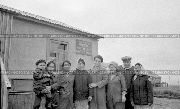 Коллективный портрет мужчины, ребенка и нескольких женщин позирующих стоя рядом с зданием, сотрудники птицефабрики под открытым небом, Лорино, Чукотка, 1970. Редакционное фото, фотограф Варфоломей Тетерин / Фотобанк Лори