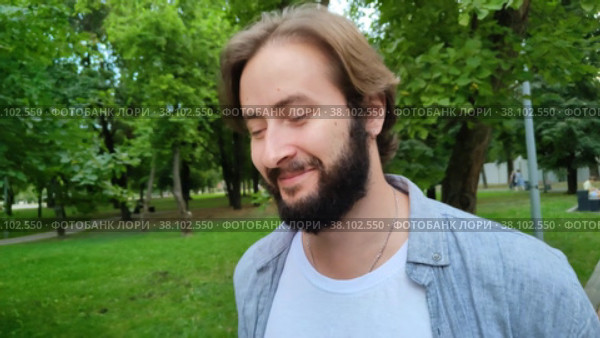 Красивый молодой мужчина идет по городскому общественному парку и улыбается. Счастливый парень гуляет по лесу или саду. Мужчина радуется теплому летнему дню. Застенчивая улыбка на лице симпатичного парня. Портрет, крупный план. Handsome young caucasian man walks through the city public park and smiles. Happy guy walks in the forest or garden. Man enjoys a warm summer day. Shy smile on the face of cute guy. Portrait, close-up. Стоковое видео, видеограф Ирина / Фотобанк Лори