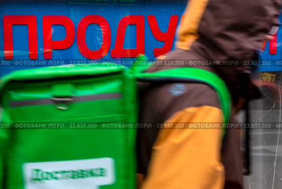 Курьер сервиса доставки еды "Яндекс Еда" идет по городу на фоне витрины продуктового магазина в центре города Москвы, Россия (2020 год). Редакционное фото, фотограф Николай Винокуров / Фотобанк Лори
