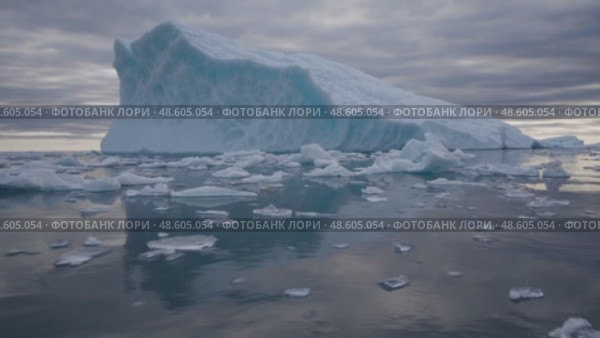 Large iceberg surrounded by drifting ice pieces in a peaceful polar seascape, under a dramatic, cloudy sky during twilight. Majestic Arctic Iceberg Floating in Calm Ocean. Стоковое AI-видео, аниматор Башкирова Анна / Фотобанк Лори
