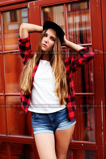 Lifestyle portrait of fashionable blonde girl with ombre hair wearing a rock red shirt, black cap, white t-shirt and denim shorts having fun outdoors in the city. Swag style. Стоковое фото, агентство Ingram Publishing / Фотобанк Лори