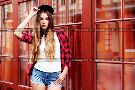 Lifestyle portrait of fashionable blonde girl with ombre hair wearing a rock red shirt, black cap, white t-shirt and denim shorts having fun outdoors in the city. Swag style. Стоковое фото, агентство Ingram Publishing / Фотобанк Лори