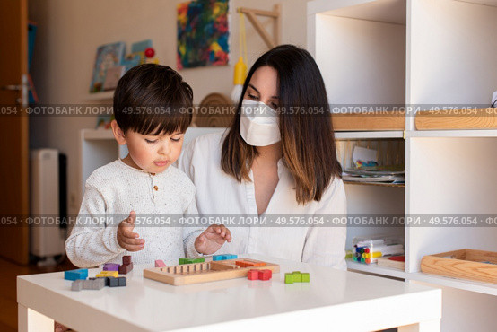 Liltle Preschooler Kid playing tetris wood puzzle with teacher educador help using face mask for coronavirus pandemic. Homeshooling. Learning Community. Montessori School. Стоковое фото, агентство Ingram Publishing / Фотобанк Лори