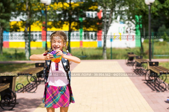 Little girl with a backpack and in a school uniform in the school yard plays pop it toy. Back to school, September 1. The pupil relaxes after lessons. Primary education, elementary class for student. Стоковое фото, фотограф Ольга Симонова / Фотобанк Лори