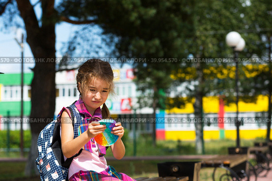 Little girl with a backpack and in a school uniform in the school yard plays pop it toy. Back to school, September 1. The pupil relaxes after lessons. Primary education, elementary class for student. Стоковое фото, фотограф Ольга Симонова / Фотобанк Лори