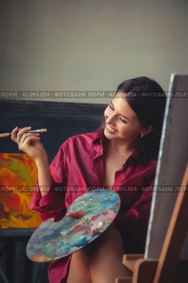 Lovely lady art painter with palette and brush in red shirt sitting at canvas, laughing. Happy perfect drawing woman artist painting in home art lab. Artisanal lifestyle concept. Copy ad text space. Стоковое фото, фотограф Aleksandr Golubev / Фотобанк Лори