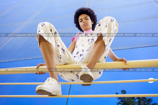Low angle full body of confident African American female in overall looking at camera while sitting on fence with bent legs. Black woman sitting on railing in city. Стоковое фото, агентство Ingram Publishing / Фотобанк Лори