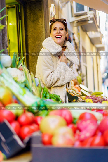 Low angle of cheerful young woman in warm clothes, smiling and looking at camera while standing near stall with bunch of fresh ripe fruits and vegetables in daylight. Gleeful young woman in cozy coat standing by fruits in shop. Стоковое фото, агентство Ingram Publishing / Фотобанк Лори