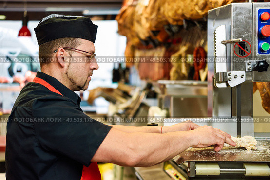 Male butcher cutting ham bones in a cutting machine inside a butcher shop.. Male butcher cutting ham bones in a cutting machine. Стоковое фото, агентство Ingram Publishing / Фотобанк Лори