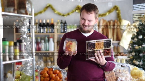 Man holds a set of dried fruit next to the shelves of food in a Christmas supermarket. Buyer chooses sweet desserts for the New Year's dinner and holiday. Стоковое видео, видеограф Яков Филимонов / Фотобанк Лори