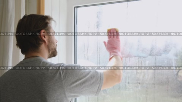 Man is cleaning window in apartment. Adult Male cleaner using a squeegee to wash a window. Bearded guy wiping and washing glasses with with spray bottle, rag and vacoom. Everyday chores. Purification. Стоковое видео, видеограф Марина Демидюк / Фотобанк Лори