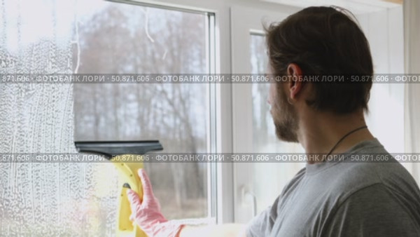Man is cleaning window in apartment. Adult Male cleaner using a squeegee to wash a window. Bearded guy wiping and washing glasses with with spray bottle, rag and vacoom. Everyday chores. Purification. Стоковое видео, видеограф Марина Демидюк / Фотобанк Лори