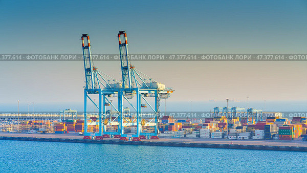 Manama, Bahrain - Jan 08 2024, Panoramic view of Bahrain commercial port with blue cranes, buildings and containers, with sea on foreground, by the morning, dawn, sunrise, Manama, Bahrain. Редакционное фото, фотограф Сергей Богомяко / Фотобанк Лори