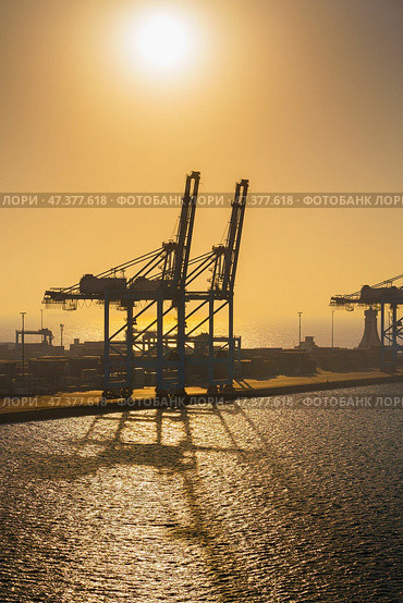 Manama, Bahrain - Jan 08 2024, Panoramic view of Bahrain commercial port with blue cranes, buildings and containers, with sea on foreground, by the morning, dawn, sunrise, Manama, Bahrain. Стоковое фото, фотограф Сергей Богомяко / Фотобанк Лори