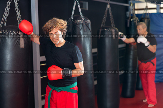 Martial artist in boxing gloves resting against punchbag in dojo. Стоковое фото, фотограф Яков Филимонов / Фотобанк Лори