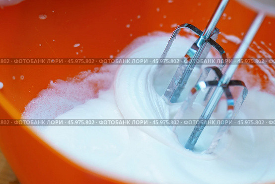 Metal mixer whisks beating a translucent egg white into a stiff white foam, set against a vibrant orange bowl, showcasing the process of baking and food preparation. Стоковое фото, фотограф Татьяна Степанищева / Фотобанк Лори