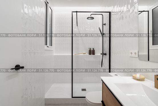 Modern bathroom with a glass shower, white tiles, and wooden accents. Minimalist design featuring a sink and decorative items on the counter. Стоковое фото, фотограф PAv / Фотобанк Лори