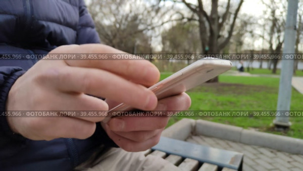 Молодой человек сидит на скамейке в городском парке, набирает текстовое сообщение или устанавливает приложение в свой мобильный телефон. Смартфон в мужских руках. Общение в социальных сетях и мессенджерах. A young man is sitting on a bench in a city park, typing a text message or installing an application in his mobile phone. The smartphone is in men's hands. Communication in social networks and messengers. Стоковое видео, видеограф Ирина / Фотобанк Лори