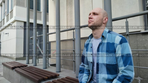 Молодой человек сидит на скамейке возле офиса или жилого здания. Задумчивый лысый парень в клетчатой рубашке. Красивый мужчина мечтает или думает о решении проблем. Человек думает о достижении своей цели. Young man sitting on bench near office or residential building. Brooding bald guy in a plaid shirt. Handsome man dreams or thinks about solving problems. Person thinks about achieving his goal. Стоковое видео, видеограф Ирина / Фотобанк Лори