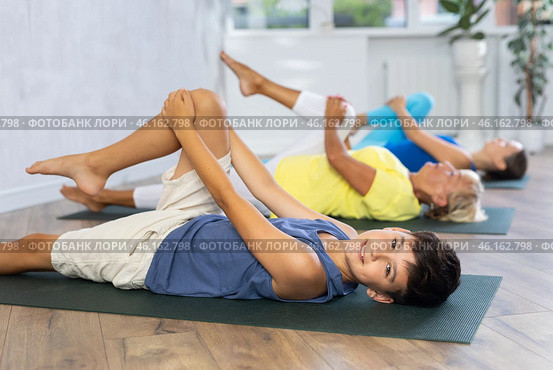 Mom, grandmother and teen grandson do preparatory stretching, lying on back and benting leg at knee. Стоковое фото, фотограф Яков Филимонов / Фотобанк Лори