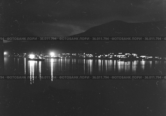 Морской пейзаж в тёмное время суток, свет от морских судов и прибрежных зданий отражается в воде, очертания холмов виднеются на противоположном берегу, Чукотка. Стоковое фото, фотограф Варфоломей Тетерин / Фотобанк Лори