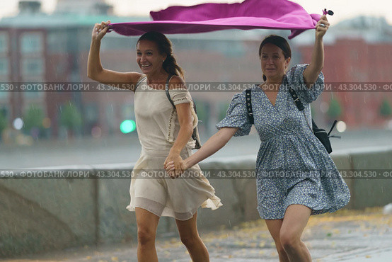 Moscow, Russia - July 10, 2022: Rainy day in the Moscow city at summer. Two young wet pretty  women runs under towel on the city street. Texture of strong, fresh and powerful water drops and sprays. Редакционное фото, фотограф YuryKara / Фотобанк Лори