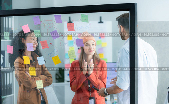 Multiethnic young business people brainstorming on a project by writing on the glass wall in the office. Стоковое фото, агентство Ingram Publishing / Фотобанк Лори