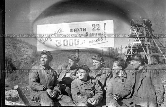 Несколько горняков позируют стоя неподалеку от каменного карьера, шахтёры жмурят глаза на солнце и смотрят в сторону, плакат с обращением к рабочим на заднем плане, Эмельджак, Якутия, 1961. Редакционное фото, фотограф Варфоломей Тетерин / Фотобанк Лори