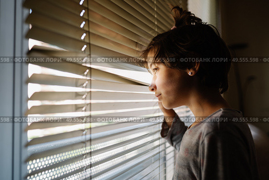 Nine-years-old girl looking out the window through the blinds in the hope of being released from quarantine due to the Covid-19 epidemic. Nine-years-old girl looking out the window through the blinds. Стоковое фото, агентство Ingram Publishing / Фотобанк Лори