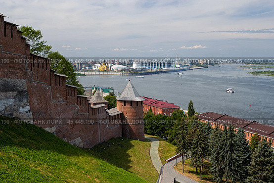 Нижний Новгород, Нижегородская область, Россия. Вид на Нижегородский кремль и Волгу. На переднем плане кирпичная крепостная стена и башни. Вдали Стрелка в месте слияния Оки и Волги. На территории Стрелки Собор Александра Невского, стадион и строительство ледовой арены. По реке плывут пассажирские и грузовые суда. (2024 год). Стоковое фото, фотограф Андрей Степанов / Фотобанк Лори