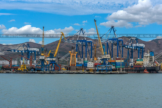 Novorossiysk Commercial Sea Port, one of the largest ports in Russia, located on the Black Sea coast. The frame shows cargo ships, port cranes, container yards and part of the industrial infrastructure. In the background are the mountain ranges surrounding the city.Numerous blue and yellow gantry cranes used for loading and unloading ships.Cargo containers stored in the port area.Commercial ships at the berths undergoing cargo operations.Tugboats used to maneuver large ships.Mountains, characteristic of the Novorossiysk landscape.Bright blue sky with clouds, creating a contrast with the industrial landscape. (2023 год). Редакционное фото, фотограф Анна Кривицкая / Фотобанк Лори