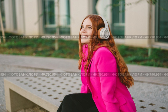 On a beautiful day, a vibrant young woman with long red hair enjoys music outdoors. Wearing stylish headphones and a bright pink jacket, she embodies carefree enjoyment and a casual lifestyle. A Young Woman Is Happily Enjoying Music While Wearing a Stylish Pink Jacket Outdoors. Стоковое фото, агентство Ingram Publishing / Фотобанк Лори