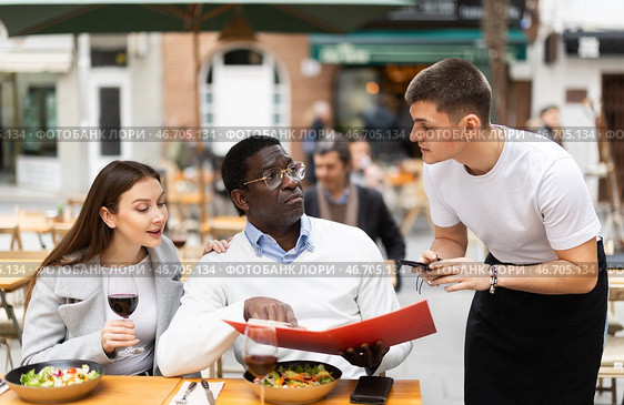 On open area of cafe, guy waiter communicates with multiethnic couple of customer, accepts order. Стоковое фото, фотограф Яков Филимонов / Фотобанк Лори