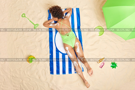 Overhead shot of relaxed slim woman dressed in green swimsuit lies on stomach sunbathes at beach applies skin care cream has lazy day enjoys summer vacation. People leisure relaxation concept. Стоковое фото, агентство Ingram Publishing / Фотобанк Лори