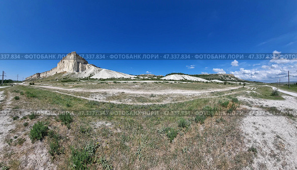 Panoramic view of the White Rock (Ak-Kaya) in Crimea, captured from a wide steppe plain. On the left rises the main part of the cliff - massive vertical walls of white limestone, as if carved by time. To the right are gentle hills and scree, forming a unique natural landscape. Blue sky, serene clouds and absolute silence emphasize the grandeur and tranquility of this place. The laid tracks from transport and thin lines of paths lead to the rock, like threads to an ancient center of power. The White Rock is not just a geological object, but a symbol of Crimean nature, filled with history, legends and power. Стоковое фото, фотограф Анна Кривицкая / Фотобанк Лори