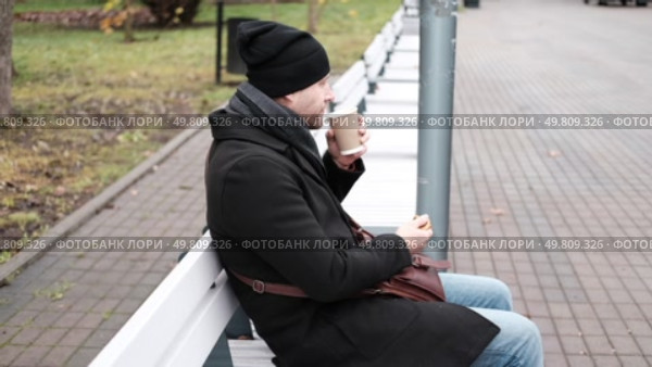 Pensive hipster drinking coffee sitting on a bench in the urban park. Handsome man wearing black coat eating street food, hot dog with paper cup in hand in the city. Stylish bearded middle-aged male. Стоковое видео, видеограф Марина Демидюк / Фотобанк Лори