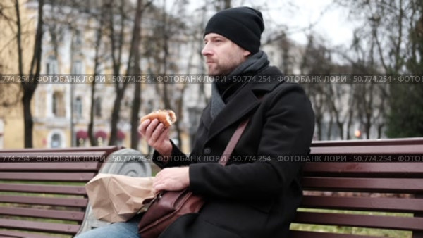 Pensive hipster drinking coffee sitting on a bench in the urban park. Handsome man wearing black coat eating street food, hot dog with paper cup in hand in the city. Stylish bearded middle-aged male. Стоковое видео, видеограф Марина Демидюк / Фотобанк Лори