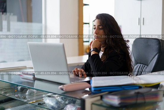 Pensive woman with wavy dark hair touching chin and browsing data on laptop while sitting at desk with sports supplies during work in gym. Thoughtful sports psychologist using netbook during work. Стоковое фото, агентство Ingram Publishing / Фотобанк Лори