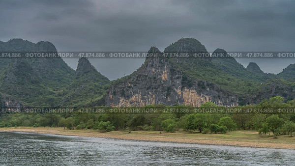 Picturesque mountains against the sky and clouds. Green vegetation on steep slopes and on the river bank. The riverbed is in the foreground. China. Li River. Li Jiang. Guilin. Стоковое фото, фотограф Вера Ивановна Кубарева / Фотобанк Лори