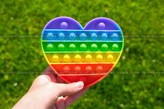 Popit - Colorful antistress sensory toy. Silicone toy - pop it in teen hand on green nature background. Rainbow color, concave and convex bubbles. Trend of summer 2021. Entertainment for children. Стоковое фото, фотограф Марина Демидюк / Фотобанк Лори