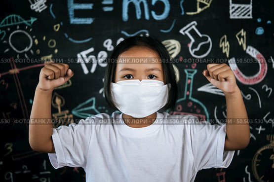 Portrait of a little girl wearing eyeglasses and facemask at school to avoid the spread of coronavirus, new normal with schoolchild,  blure blackboard in background. Стоковое фото, агентство Ingram Publishing / Фотобанк Лори