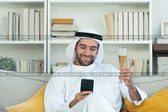 Portrait of a smiling Arabian man holding a glass of champagne and using mobile phone at home. Стоковое фото, агентство Ingram Publishing / Фотобанк Лори